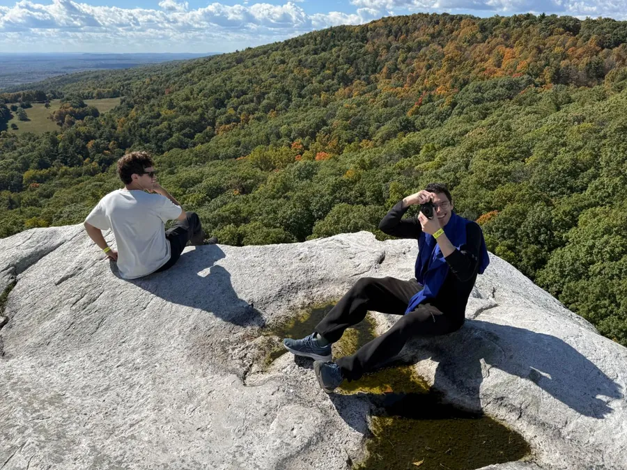 Two teammates sitting on a rock summit above fall foliage, one taking a photo